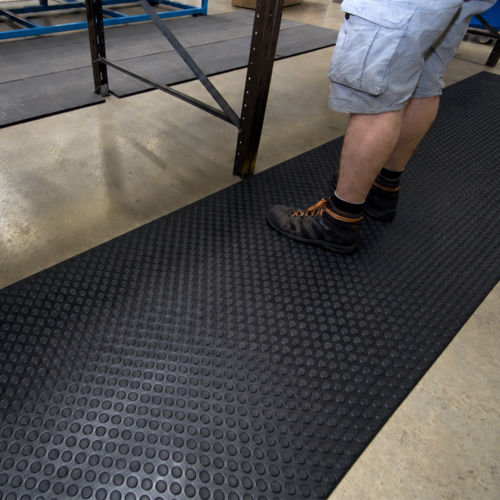 Trabajador de pie sobre una alfombra antifatiga negra Orthomat® Dot con superficie de relieve circular en un taller industrial.