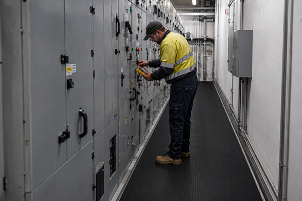 An electrician tests electrical switchboard panels while standing on black electrical safety matting in a narrow switchboard room.