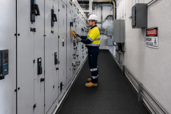 An electrician wearing PPE tests an electrical switchboard in a high voltage switchboard room, standing on black electrical safety matting.