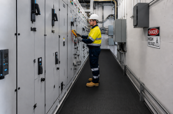 An electrician wearing PPE tests an electrical switchboard in a high voltage switchboard room, standing on black electrical safety matting.