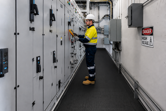 An electrician wearing PPE tests an electrical switchboard in a high voltage switchboard room, standing on black electrical safety matting.