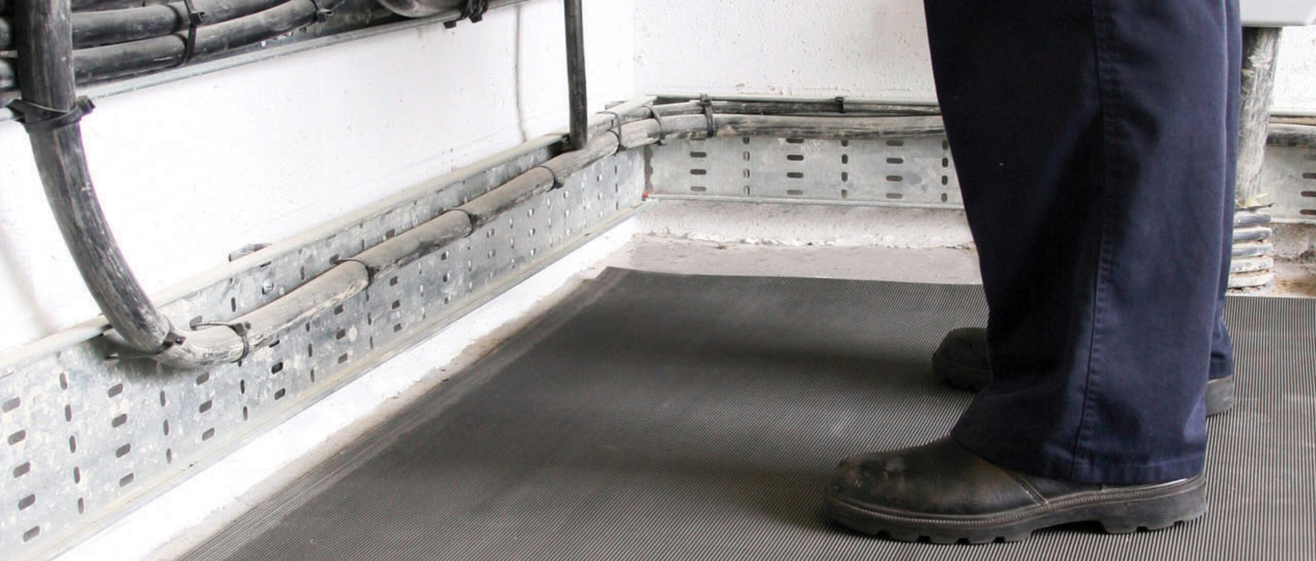 Close up of worker’s boots standing on grey electrical insulation matting near high voltage cables and switchboard equipment.