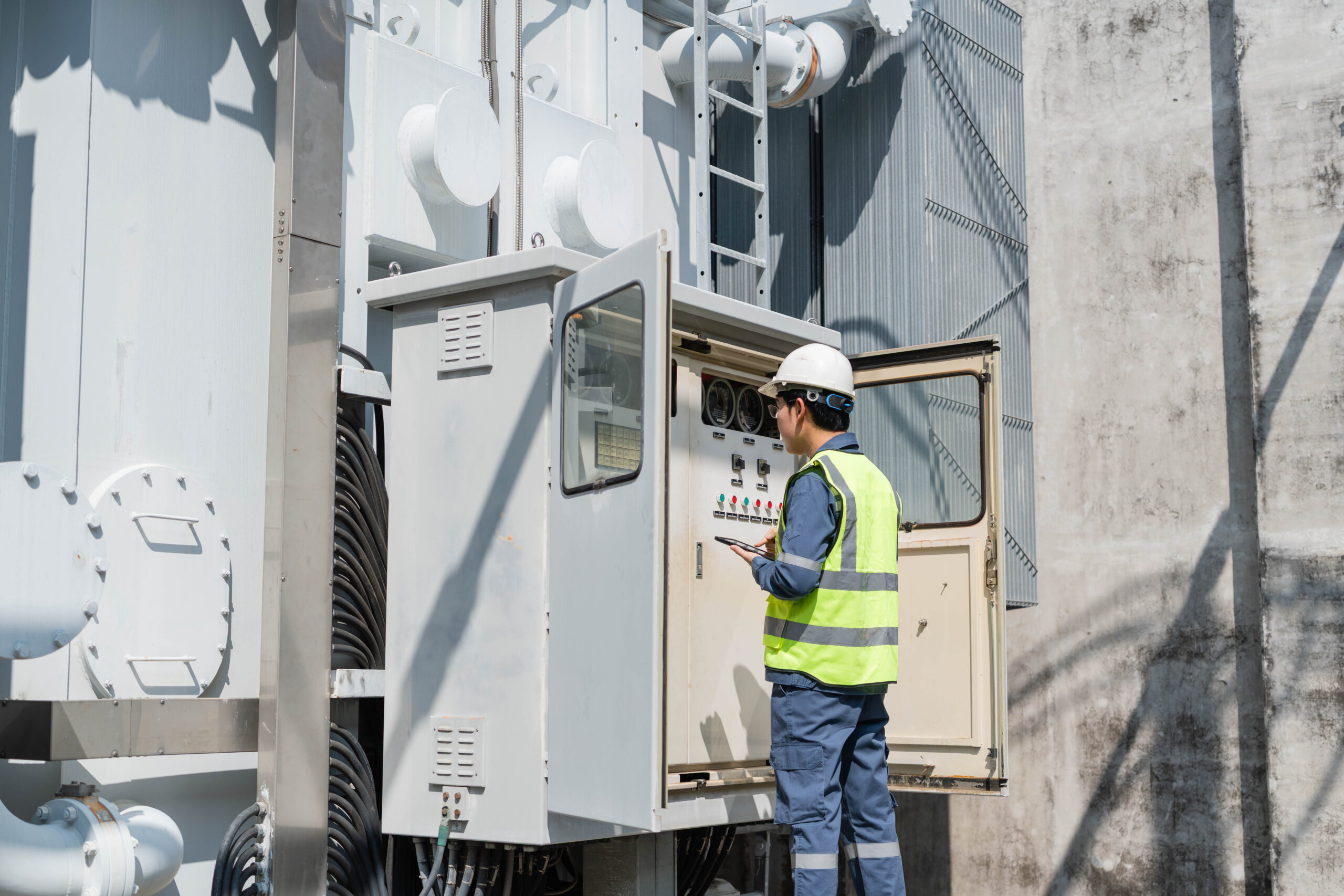 Worker inspecting electrical control equipment beside a large industrial transformer.
