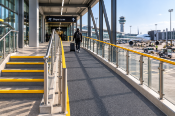Airport access ramp with dark GRP anti-slip flooring beside standard stairs fitted with yellow GRP stair nosings, with a traveller pulling wheeled luggage toward the terminal.